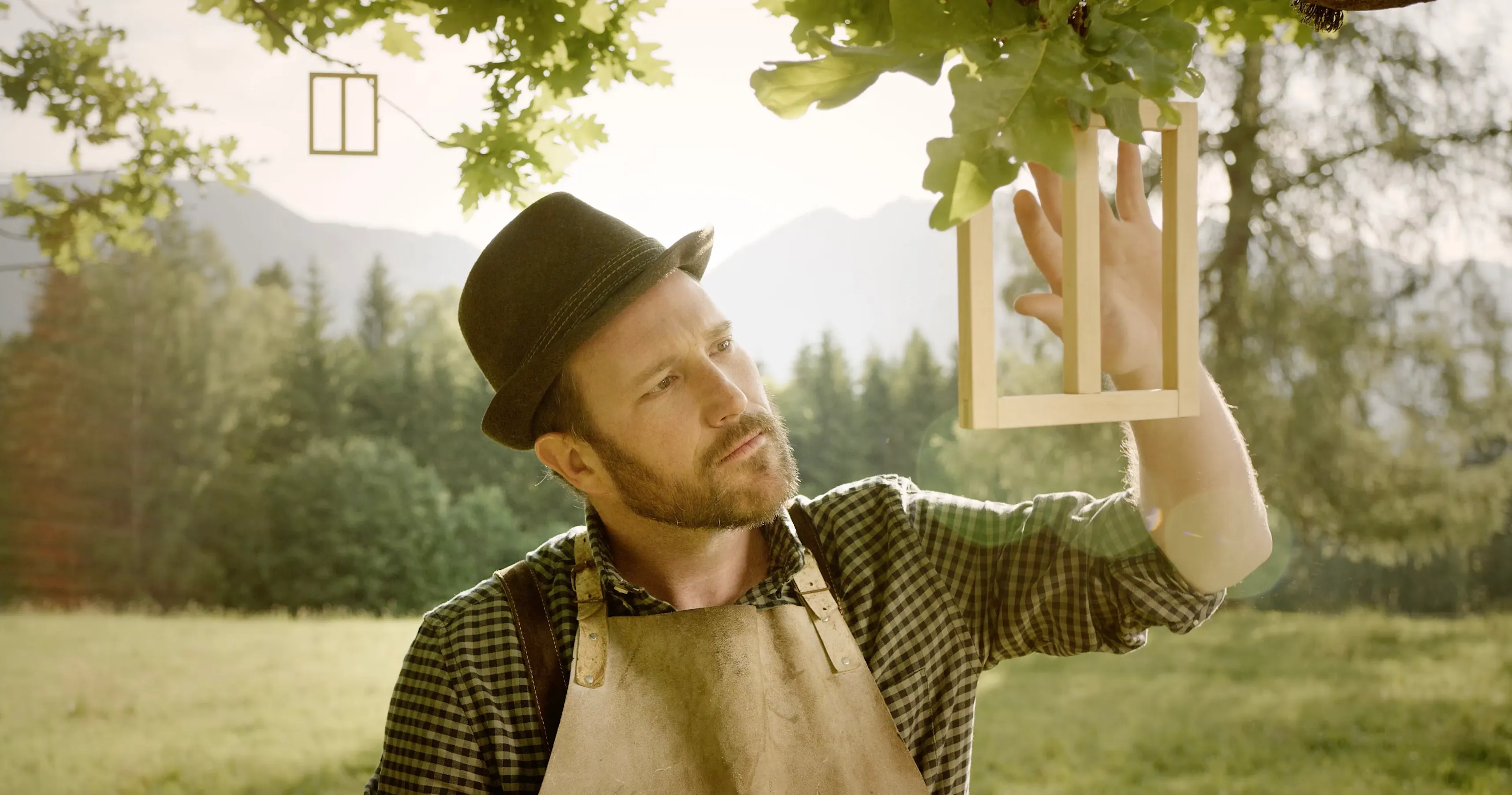 Ein Mann mit Hut und Lederschürze hält unter einem Baum einen hölzernen Fensterrahmen in die Höhe