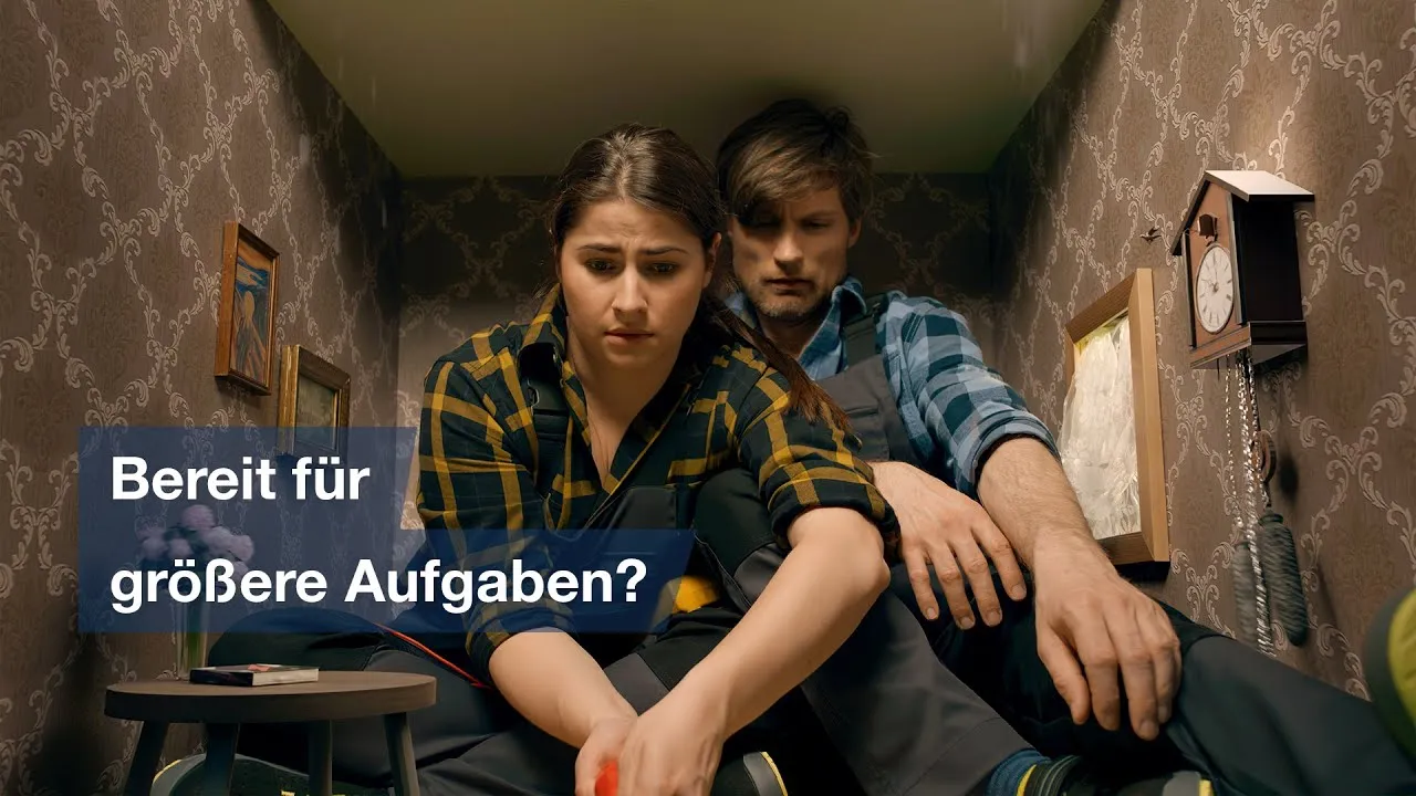 Eine Frau und ein Mann in Arbeitskleidung kauern eingezwängt in einem viel zu kleinen Zimmer mit Tapete und Kuckucksuhr