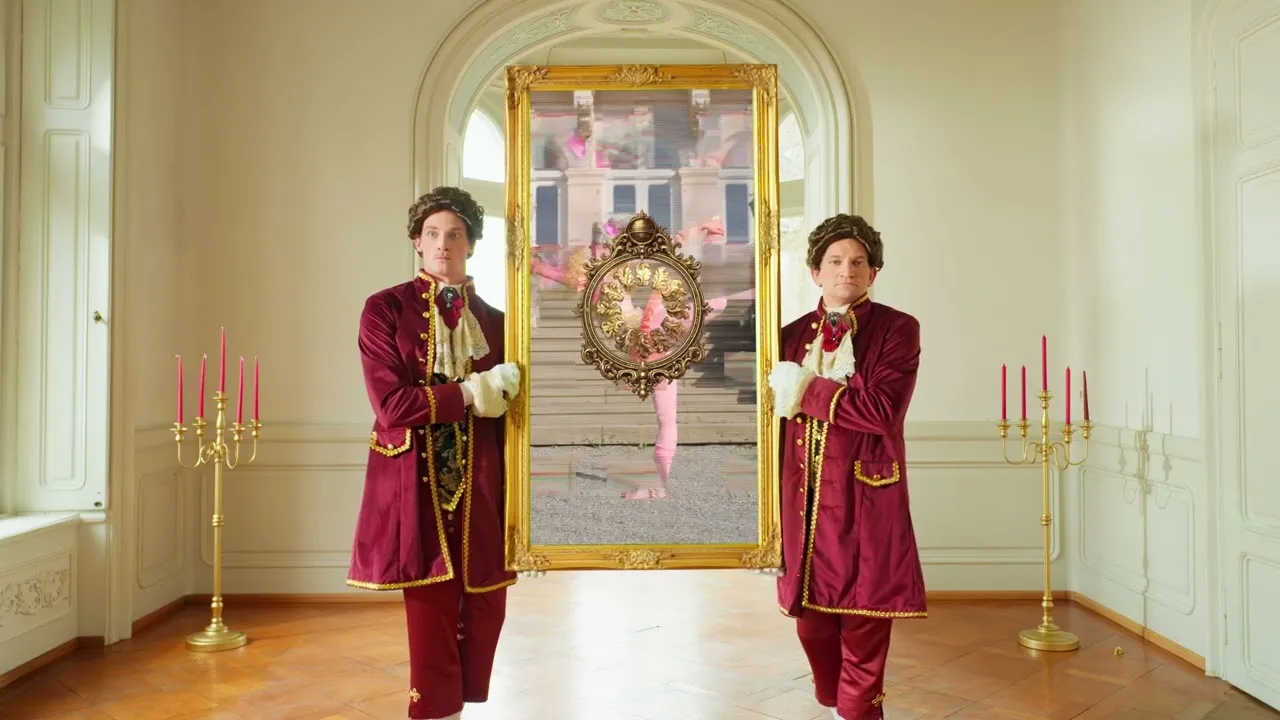Zwei Diener in barocken Samtröcken und Perücken tragen einen goldenen Spiegel durch einen Schlosssaal